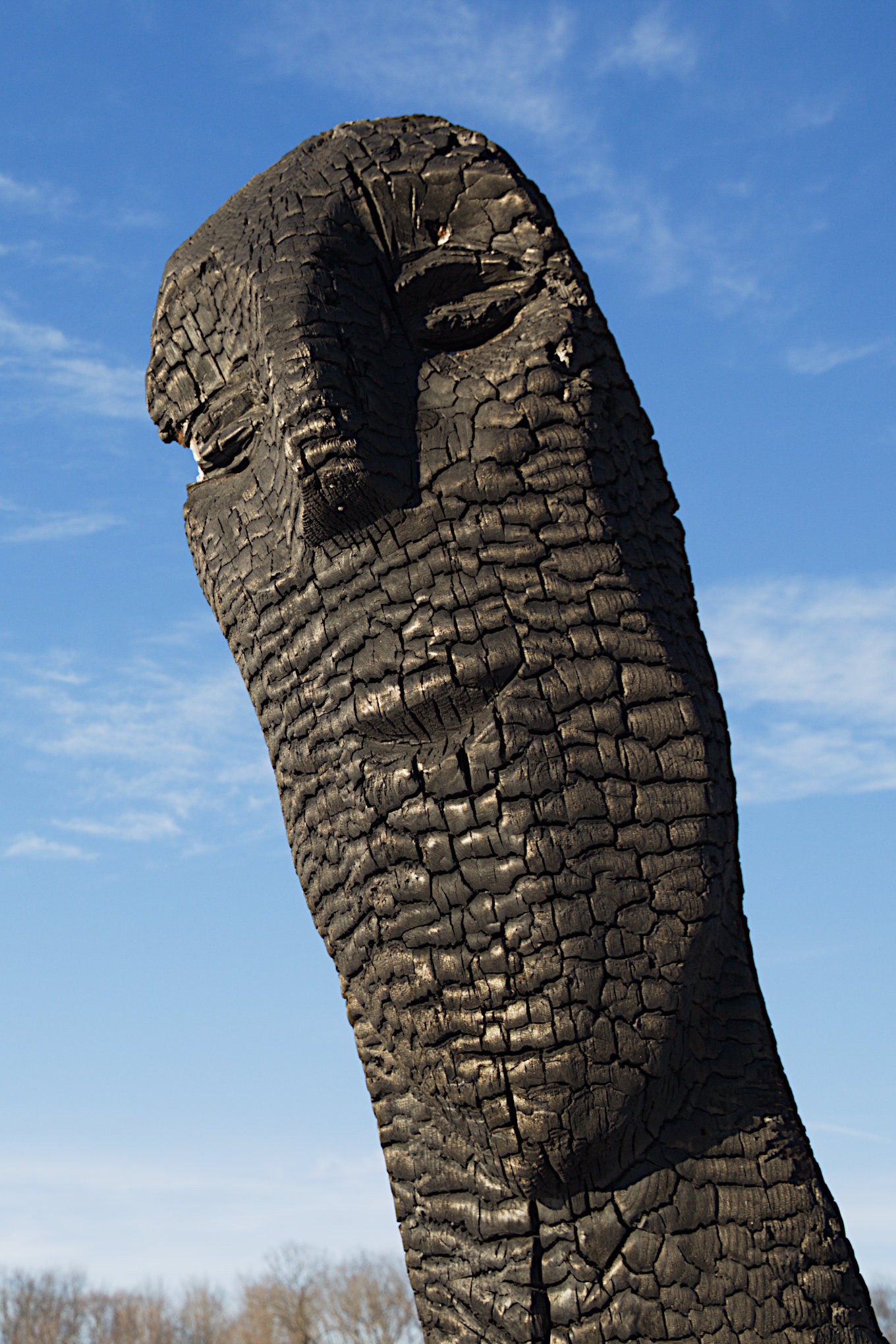 A charred carved wooden face standing against a clear blue sky