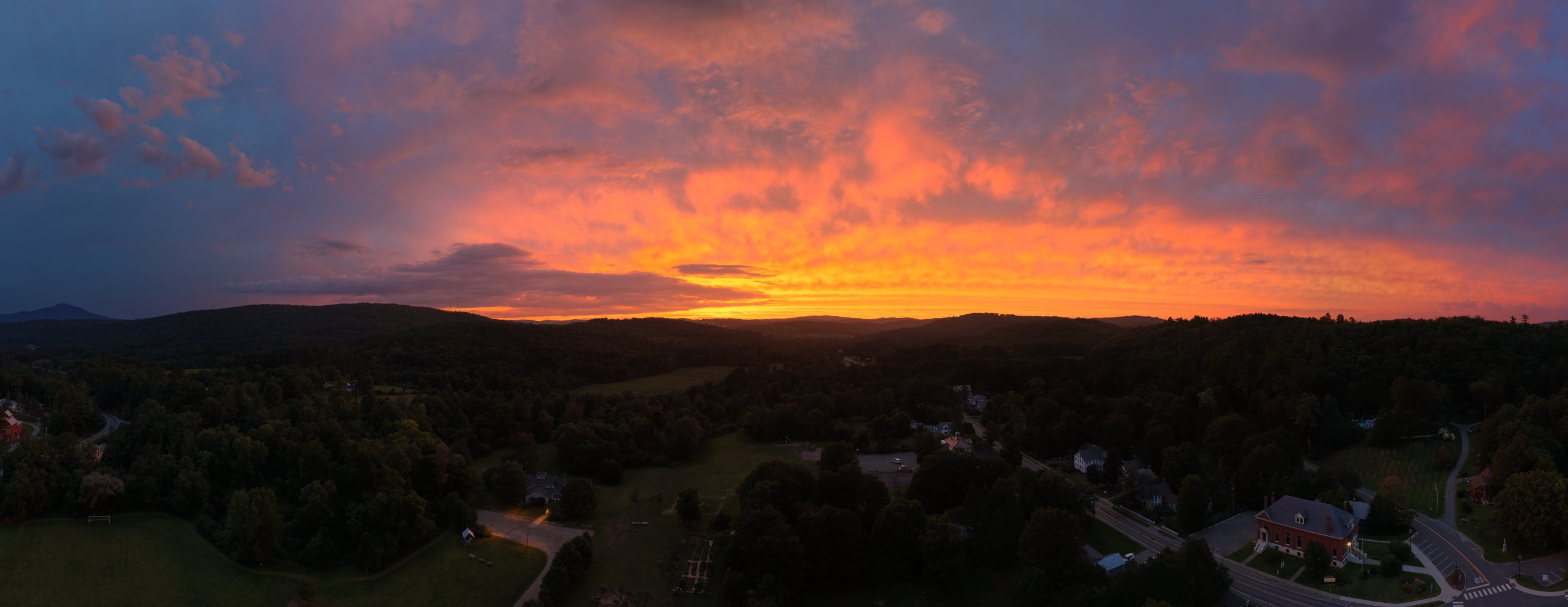 Wide panorama of Hartland, Vermont at sunset — the whole sky burning orange and violet over the village, Damon Hall lit below